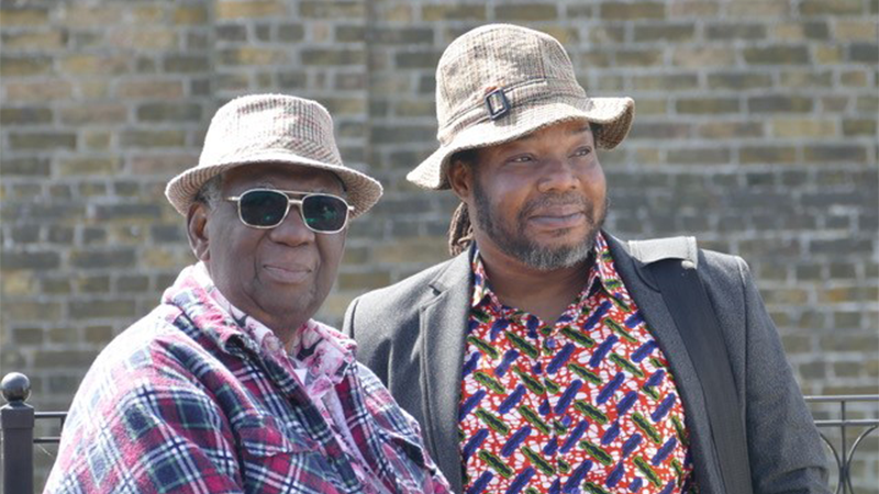 Two men stand together outdoors in casual attire, both wearing patterned hats