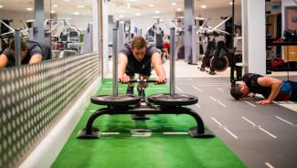 A man pushes a weighted sled on turf, while another does push-ups in a gym